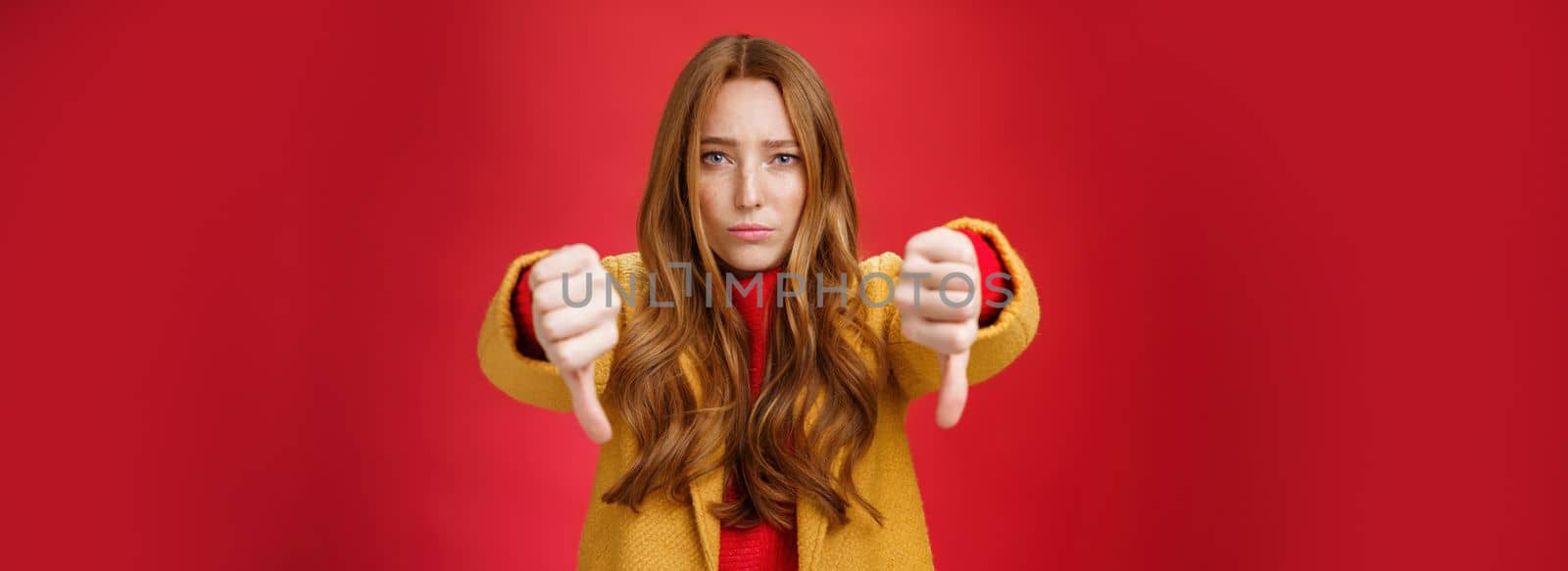 Lifestyle. Portrait of insecure and unconfident cute redhead female reacting to uncool thing showing thumbs down with worried upset expression from dislike and disappointment over red background.