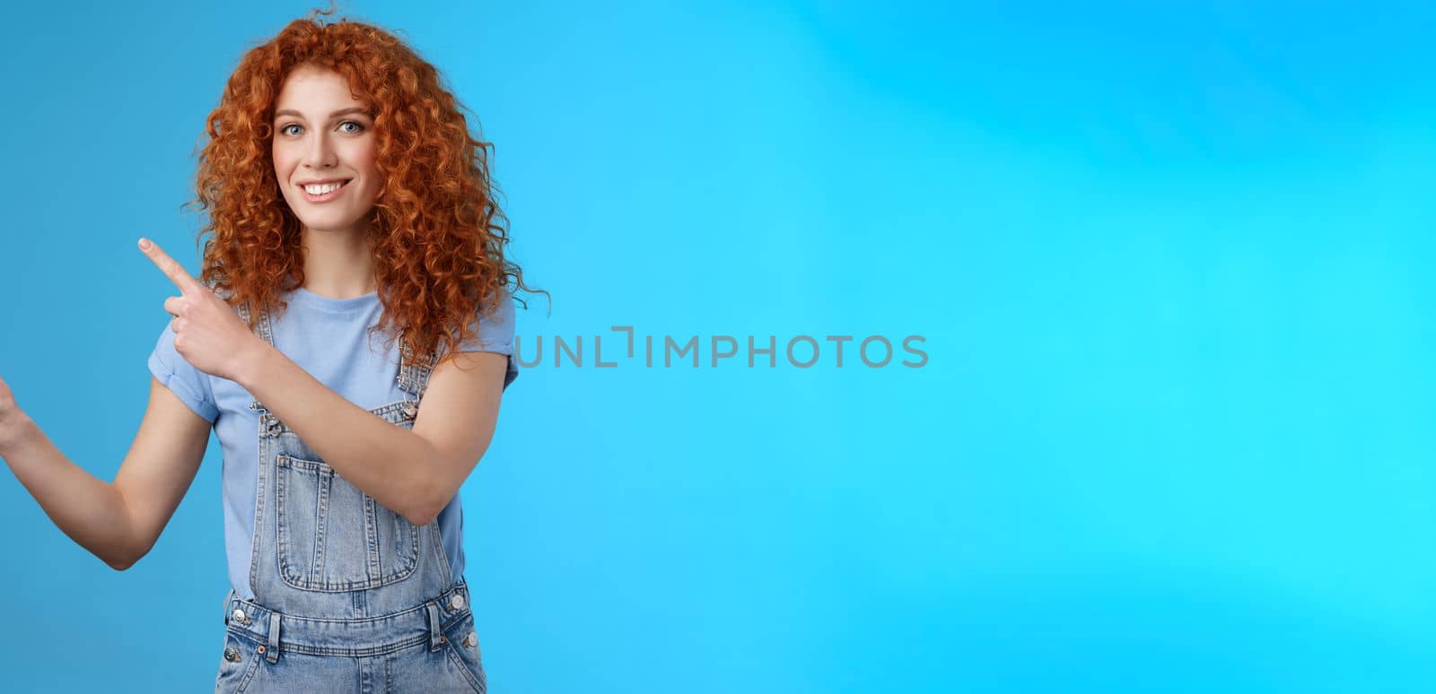 Cheerful upbeat positive summer girl redhead curly hairstyle wear denim overalls getting ready beach vacation pointing upper left corner promote store smiling delighted enjoy holidays.