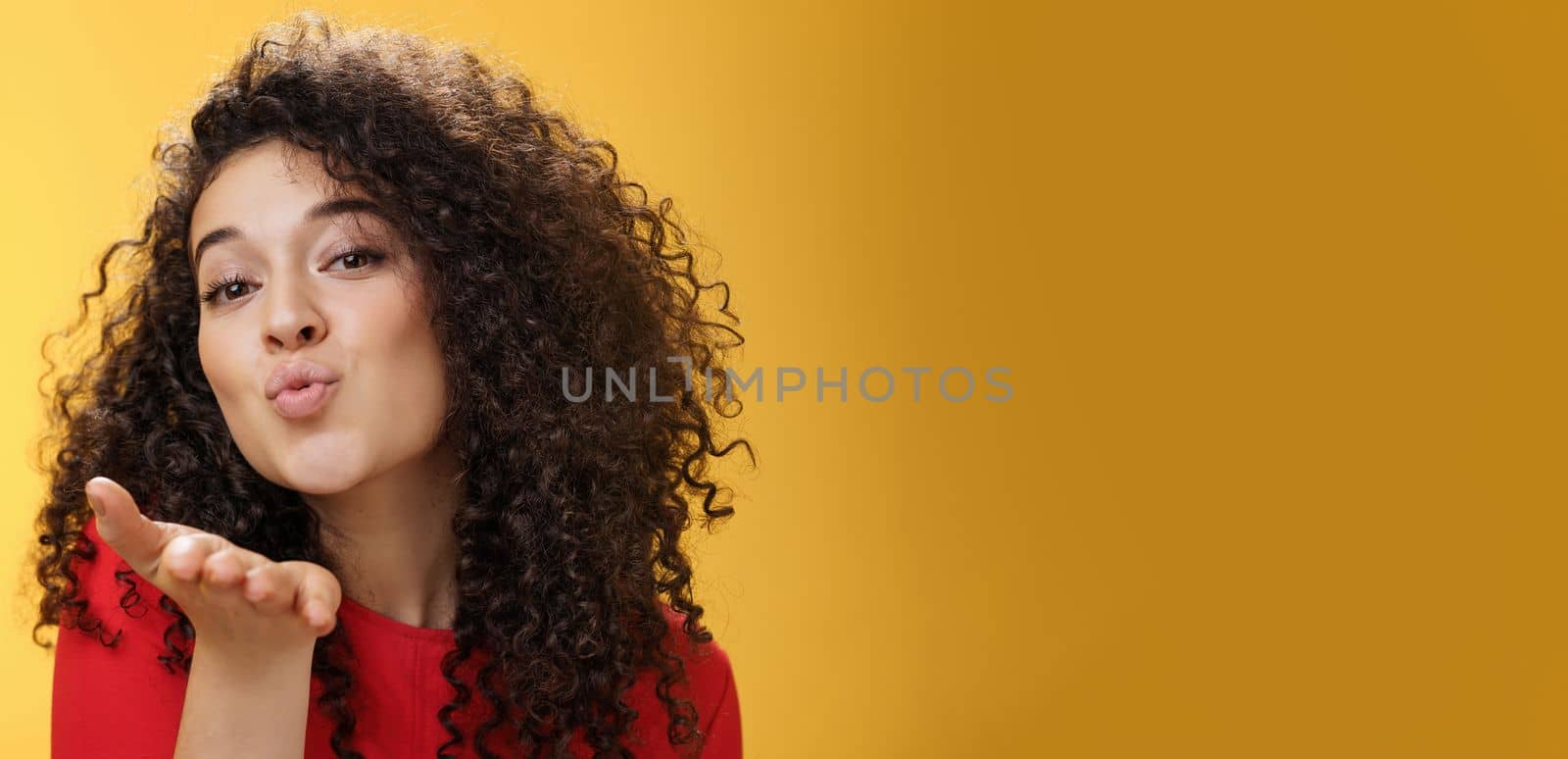 Sensual flirty and tender attractive girlfriend with curly hairstyle folding lips extending hand to send air kiss at camera, smiling flirting and being coquettish over yellow background. Romance, people and relationship concept