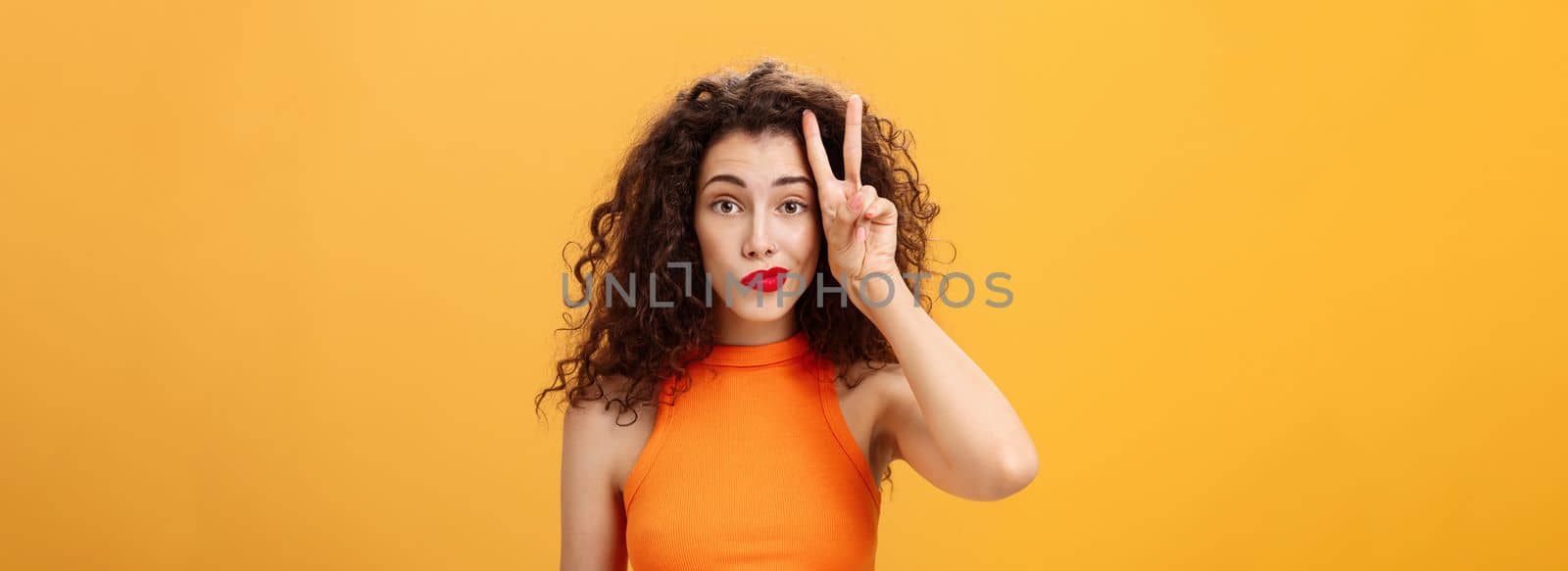 Waist-up shot of charismatic good-looking european female. with curly haircut in cropped top showing peace sign near face making silly look smirking and gazing friendly at camera over orange background.