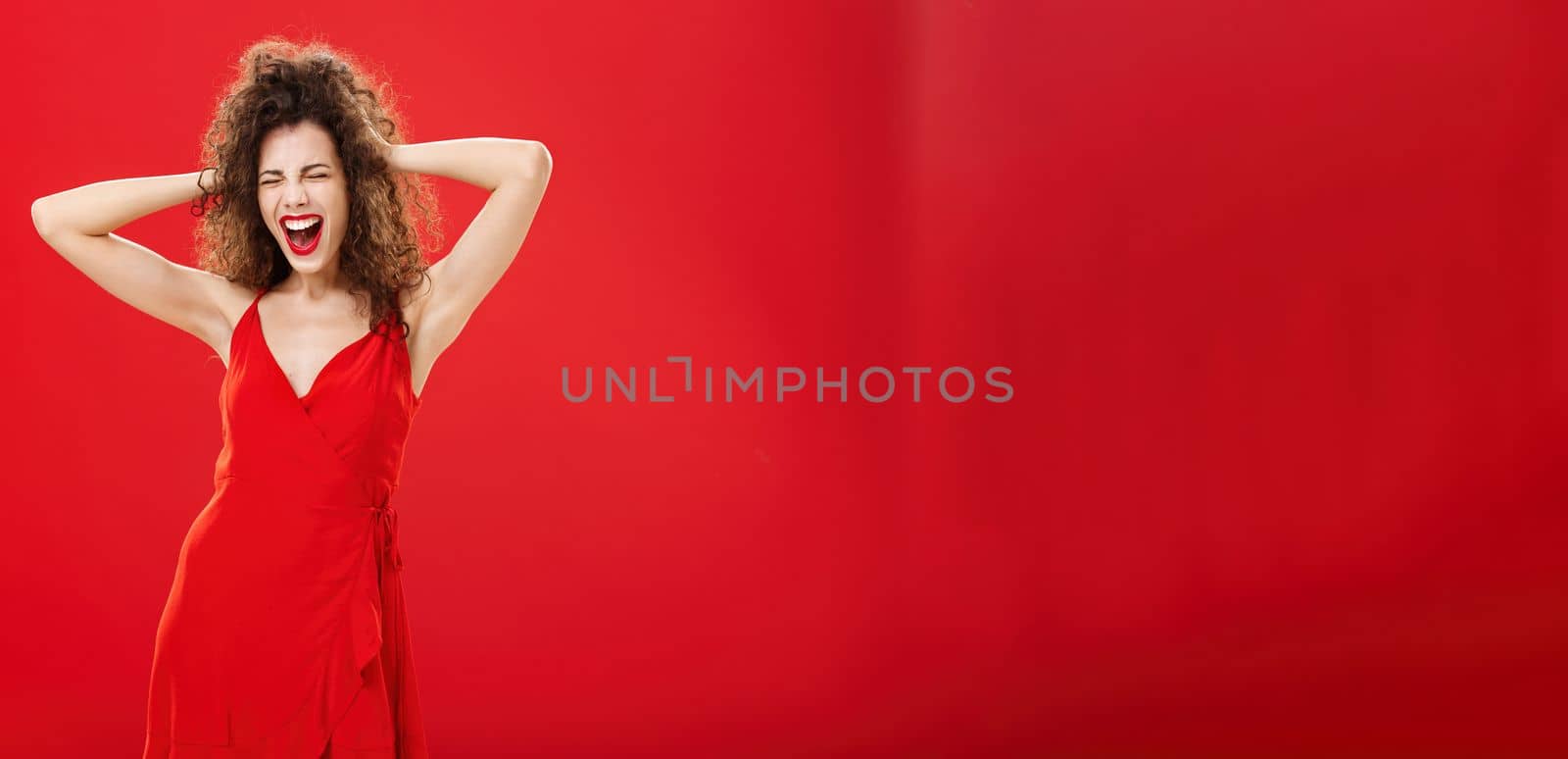 Girl just wanna have fun. Crazy adult caucasian woman with curly hairstyle in elegant dress holding hands on head yelling out loud with closed eyes and intense expression over red background.