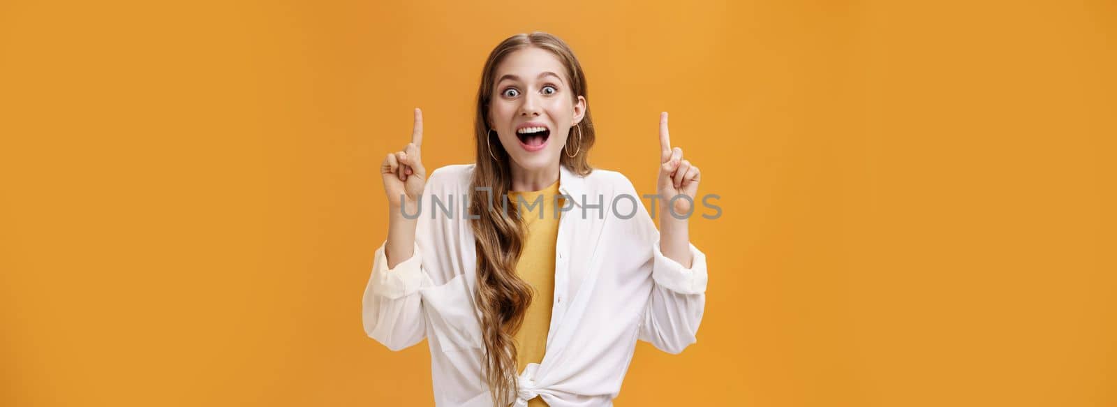 Looking up on bright side. Charismatic happy enthusiastic young attractive woman in white blouse dropping jaw with wide joyful smile pointing up with raised index fingers showing awesome thing.