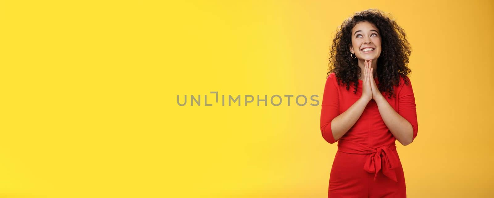 Lifestyle. Dreamy excited cute b-day girl with curly hair in cute red dress rubbing palms together near chest as hands in pray smiling looking up delighted and hopeful making wish over yellow background.