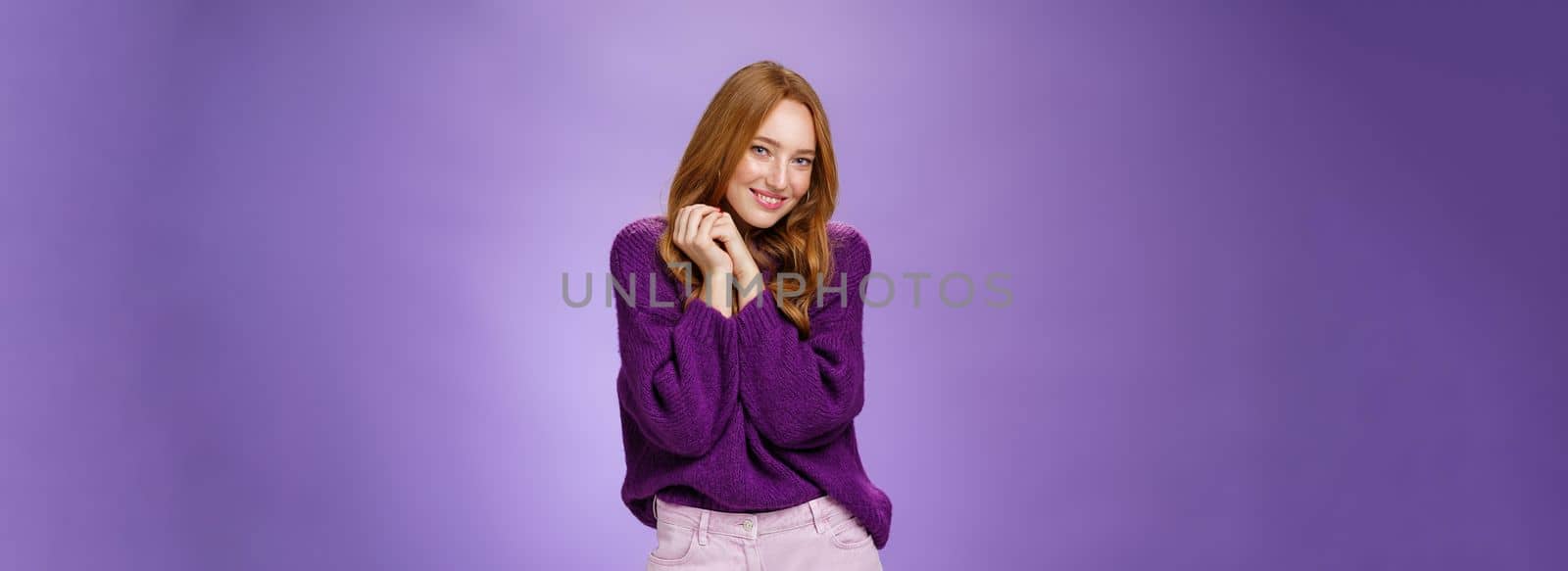 Cute and tender redhead girlfriend making romantic glances, flirting and giggling sweet holding hands together near shoulder smiling and looking at camera shy and coquettish over purple wall.