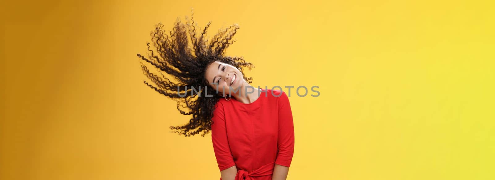 Bright happy and carefree playful woman waving curly hair making wave and smiling broadly as standing joyful in red dress over yellow background in good mood for future adventures by Benzoix