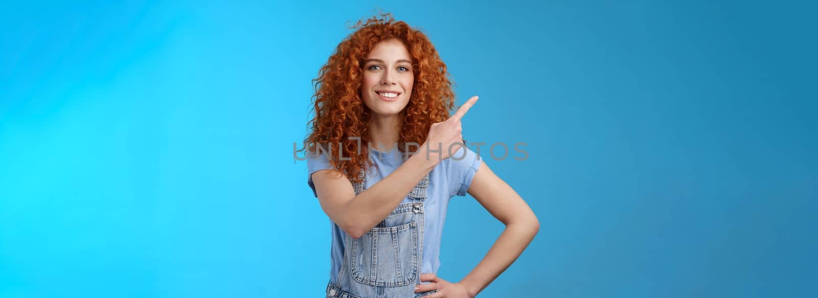 Motivated good-looking confident redhead curly girl dungarees feel empowered showing woman deal anything pointing upper left corner assertive assured you gonna like product, blue background.