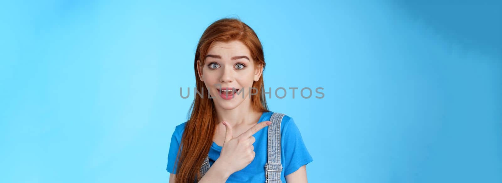 Curious excited redhead woman attend interesting exhibition, pointing left look camera interested, questioned about copy space promo, asking question intrigued cool offer, blue background.