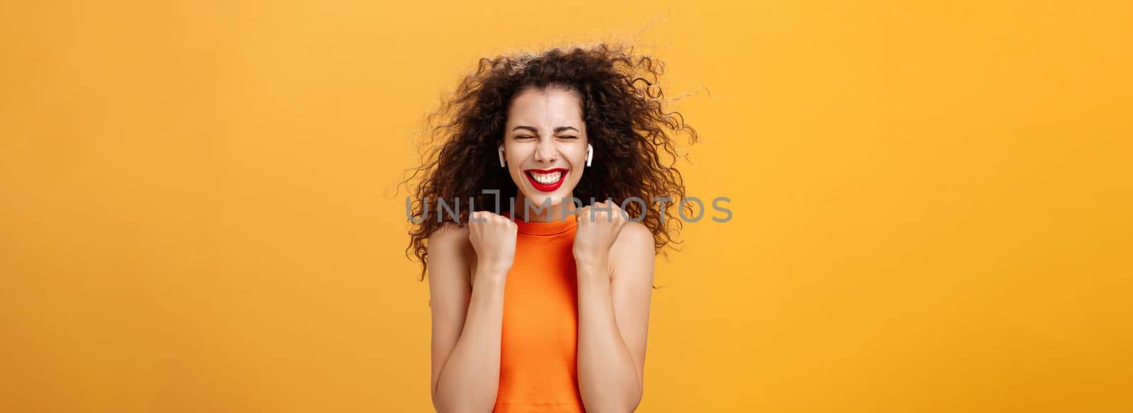 Pleased happy and triumphing curly-haired female in cropped top clenching fists in yes gesture closing eyes and smiling broadly receiving awesome news, winning award over orange background. Copy space