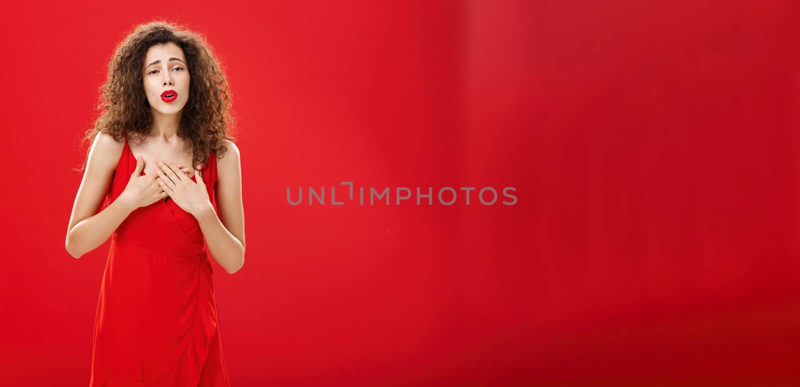 Portrait of touched elegant and stylish curly-haired adult. woman receiving compliments holding hands on chest in grateful and thankful pose hearing heartwarming speech in her honor over red background.