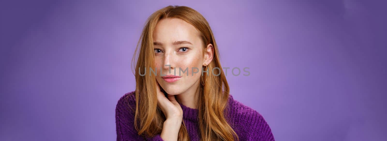 Sensual and romantic woman with red hair and freckles touching cheek gently as curls dropping on half face gazing charmed and flirty at camera, smiling being tender and coquettish over purple wall.