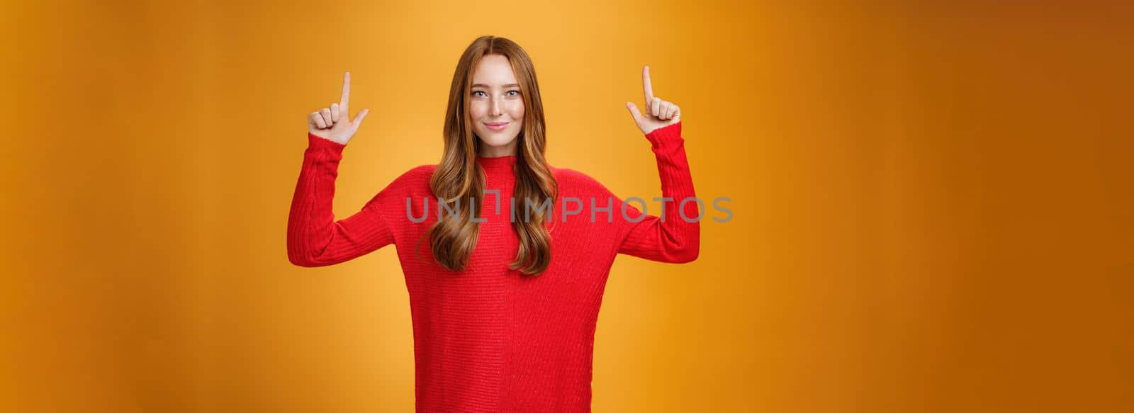 Look up you not regret it. Portrait of confident and stylish young redhead woman in 20s wearing knitted red dress pointing upwards and smiling assertive with self-assured grin over orange background.