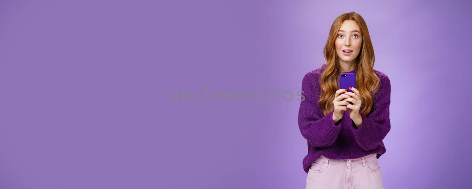 Impressed speechless cute redhead female actor receiving awesome surprising message on mobile phone open mouth from amazement and raising eyebrows astonished, posing over purple wall. Technology and people emotions concept