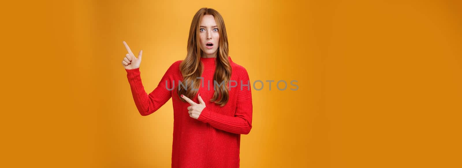 Shocked distressed and confused ginger girl with freckles and blue eyes in red sweater pointing at upper right corner with shook and surprise open mouth, raise eyebrows from stupor over orange wall.