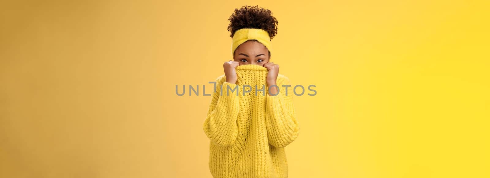 Charming cute silly young african-american modern girl. pulling sweater on face hiding peeking camera playfully having fun smiling hide-n-seek fool around mimicking standing yellow background.