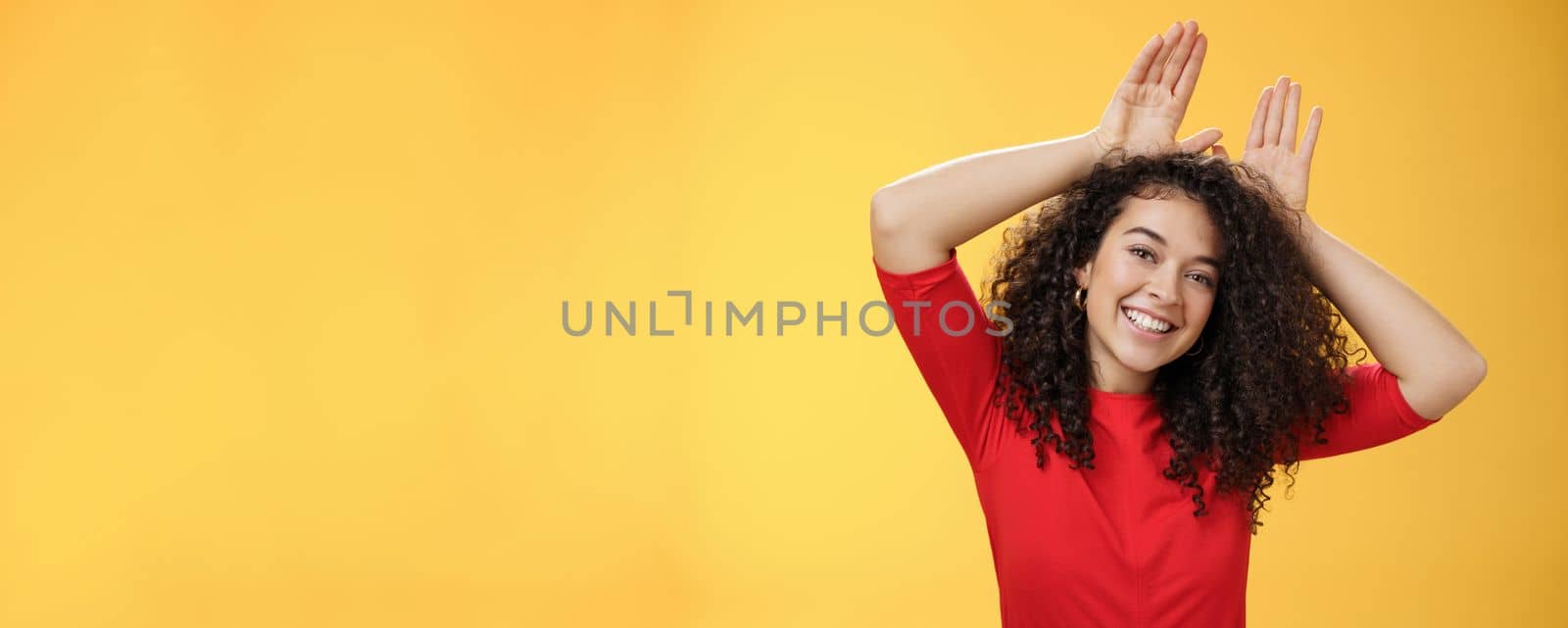 Close-up shot of charismatic playful and tender young kind woman with curly hair playing having fun showing bunny ears with hands on head tilting head joyfully fooling around and smiling at camera by Benzoix