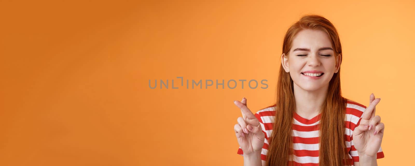 Hopeful optimistic cheerful ginger girl believe dream come true, close eyes cross fingers good luck, making wish, anticipating positive good fortune, stand orange background smiling daydreaming.