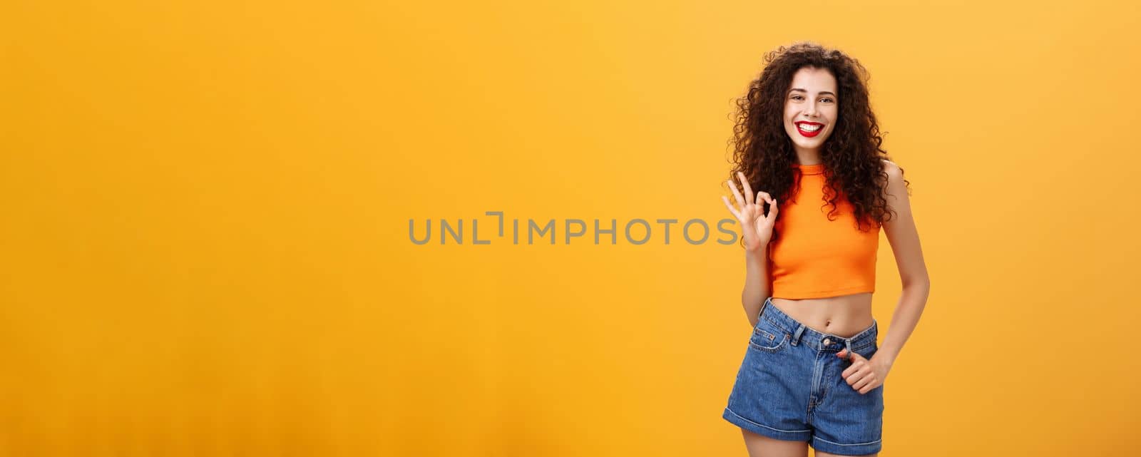 Charming young charistmatic woman. with curly hairstyle in cropped top and shorts showing okay or approval gesture smiling broadly liking awesome idea of friend where hang out over orange wall.