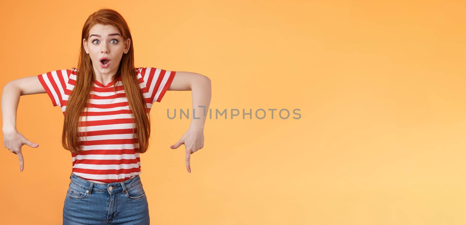 Shocked excited attractive redhead woman gasping, drop jaw shook, stare camera astonished, pointing down, feel amazed and surprised see incredible good offer, gossiping stunned, orange background.