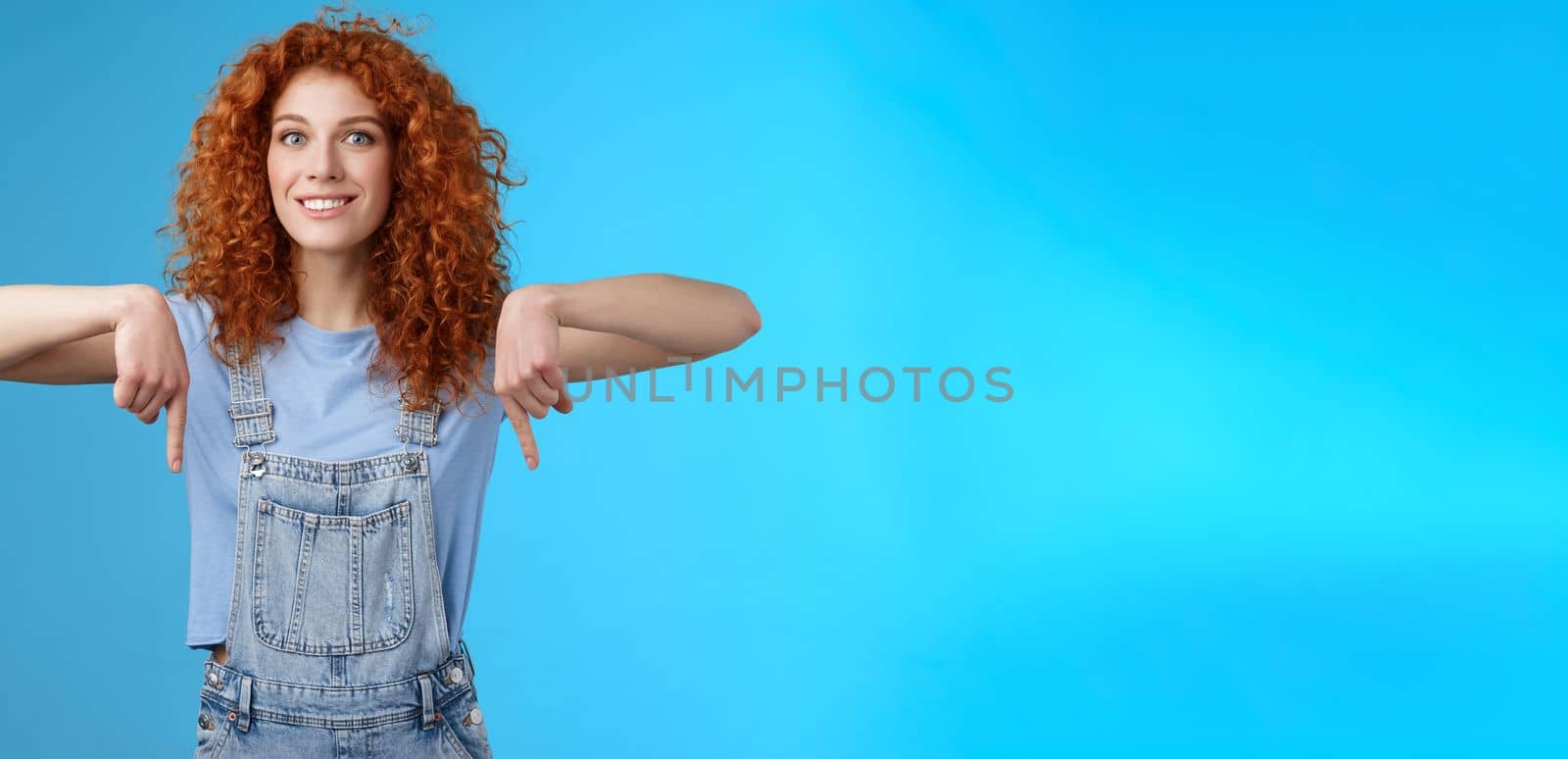 Excited happy smiling caring redhead curly-haired girlfriend thrilled awaiting summer holiday trip pointing down index finger upbeat mood show favorite store buy best prices, blue background by Benzoix