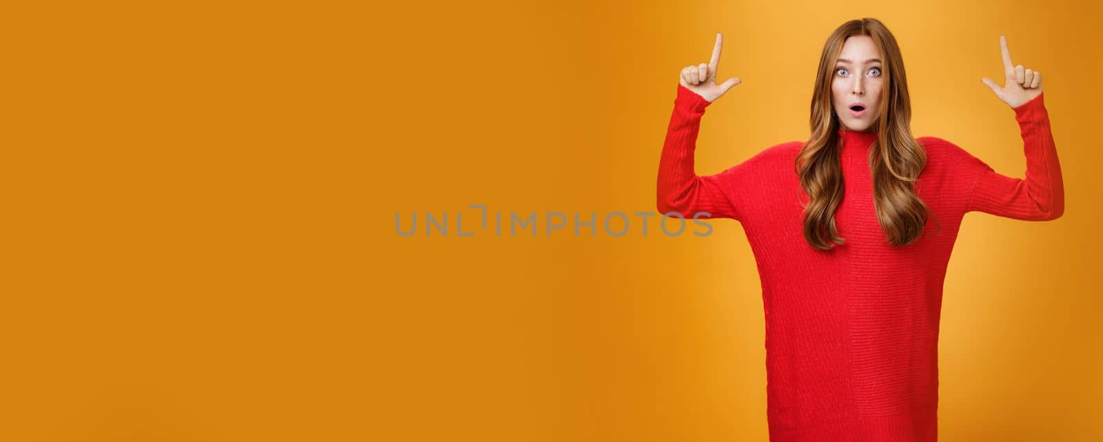 Portrait of attractive and stylish redhead 25s female in knitted red dress gasping from amazement open mouth questioned and surprised as pointing up at astonishing promotion over orange wall by Benzoix