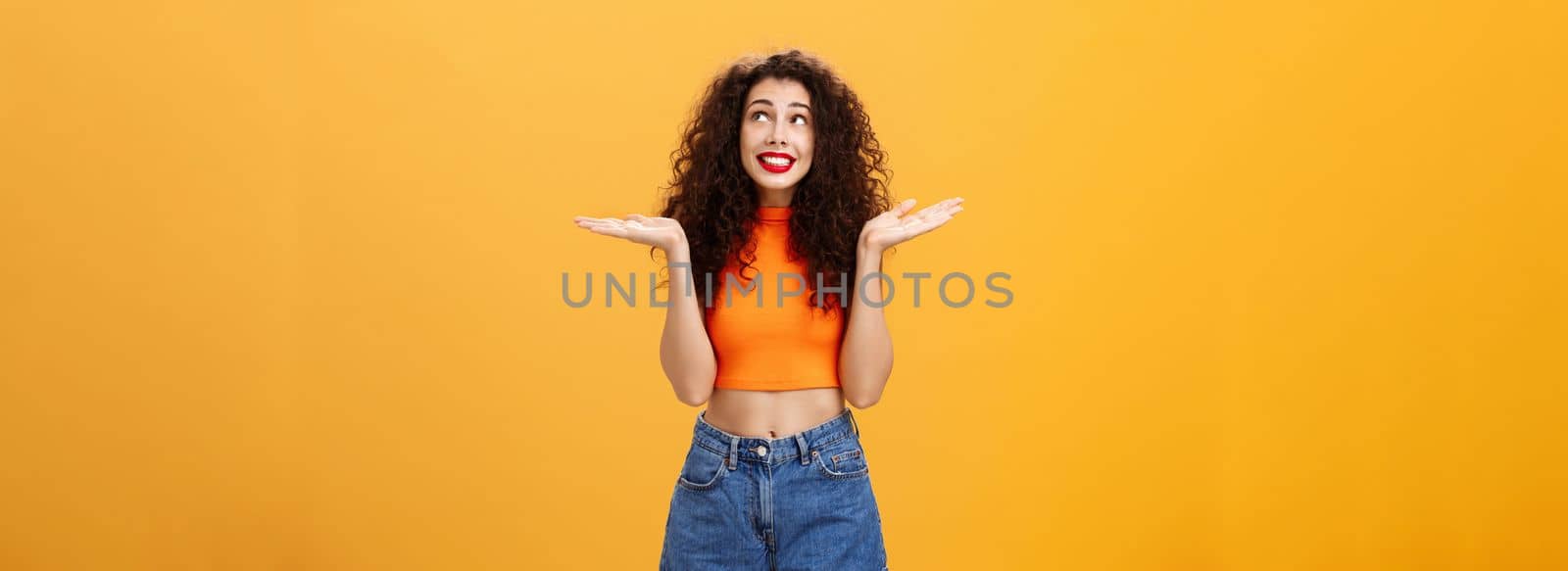 Silly and clueless carefree cute girl. with curly hairstyle in cropped top and shorts shrugging with palms spread aside smiling unaware and gazing at upper right corner uncertain and uninvolved.