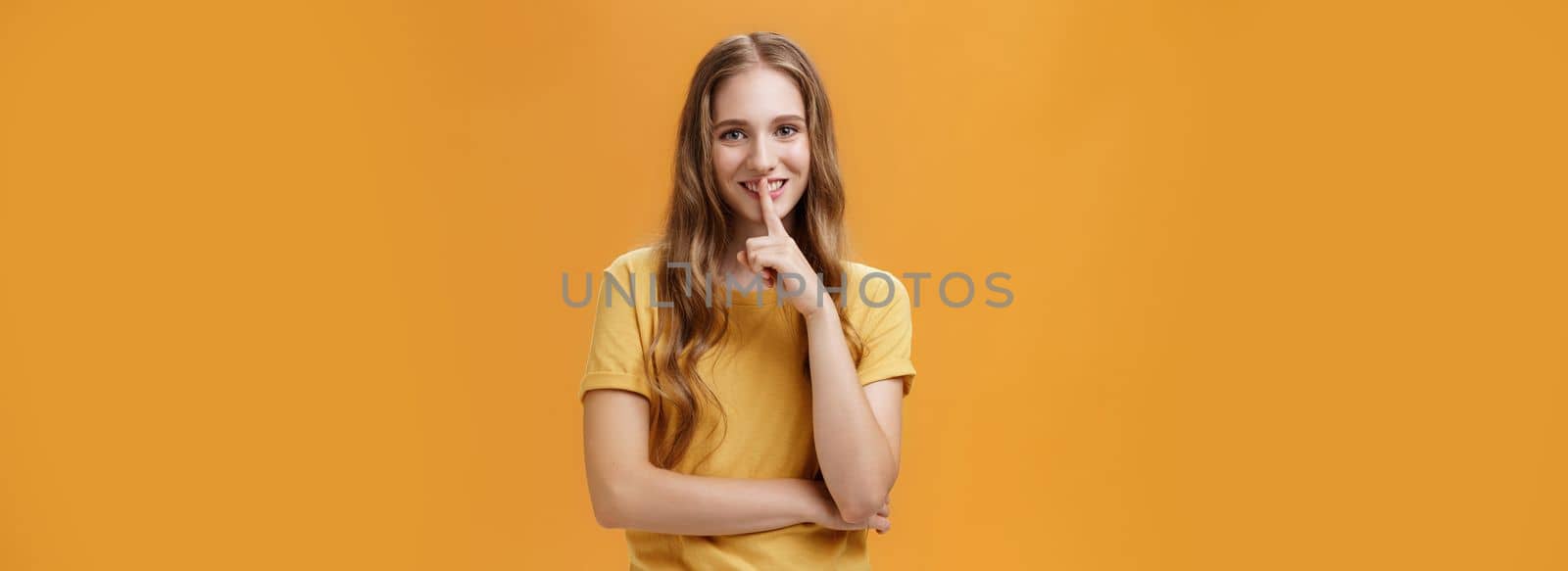 Girl wants keep secret betweet us. Portrait of charming good-looking slim pretty and young woman with long wavy hair and tattoo showing shh gesture with index finger over mouth against orange wall.