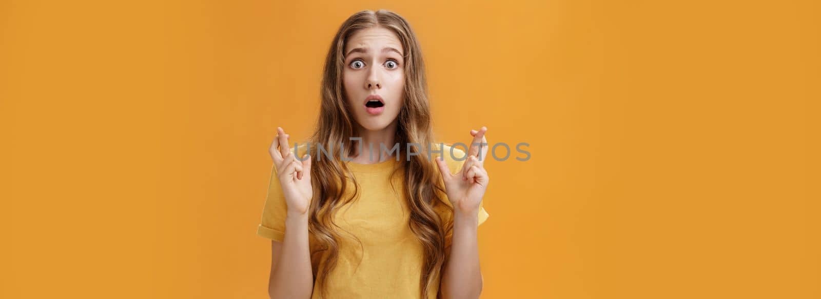 Portrait of worried silly young cute woman with wavy natural hairstyle crossing fingers for good luck opening mouth looking with hope and faith at camera praying, making wish over orange wall.