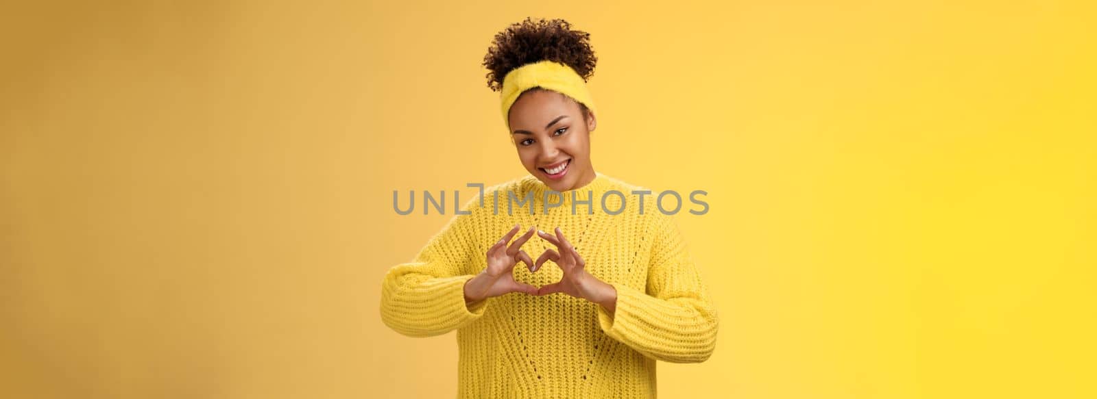 Lovely tender stylish african-american girl curly hairstyle headband sweater showing heart love gesture gladly smiling flirty tilting head express sympathy passion desire, posing yellow background.