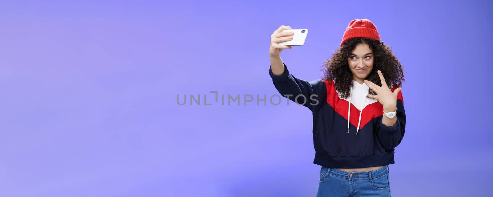 Lifestyle. Cool and stylish good-looking caucasian female with curly hair in trendy red beanie and sweatshirt looking from under forehead awesome and swah taking selfie with smartphone holded in extended arm.