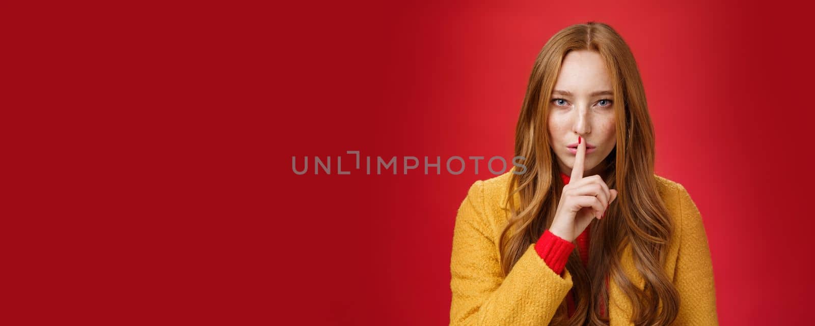 Shh keep out secret safe. Portrait of sensual attractive and daring redhead female in yellow coat showing shush gesture with index finger on lips, squinting looking determined and sassy over red wall.