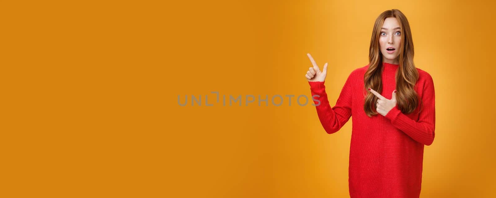 Lifestyle. Surprised and impressed intrigued nice cute redhead female with freckles in warm red sweater pointing at upper left corner open mouth from amazement and looking questioned at camera over orange wall.