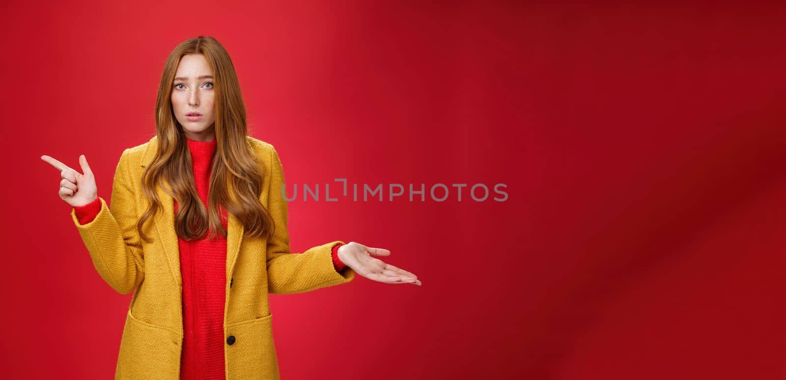 Lifestyle. Frustrated and confused young cute female with ginger hair in yellow coat shrugging as pointing questioned left waiting explanation as asking question clueless and puzzled over red background.