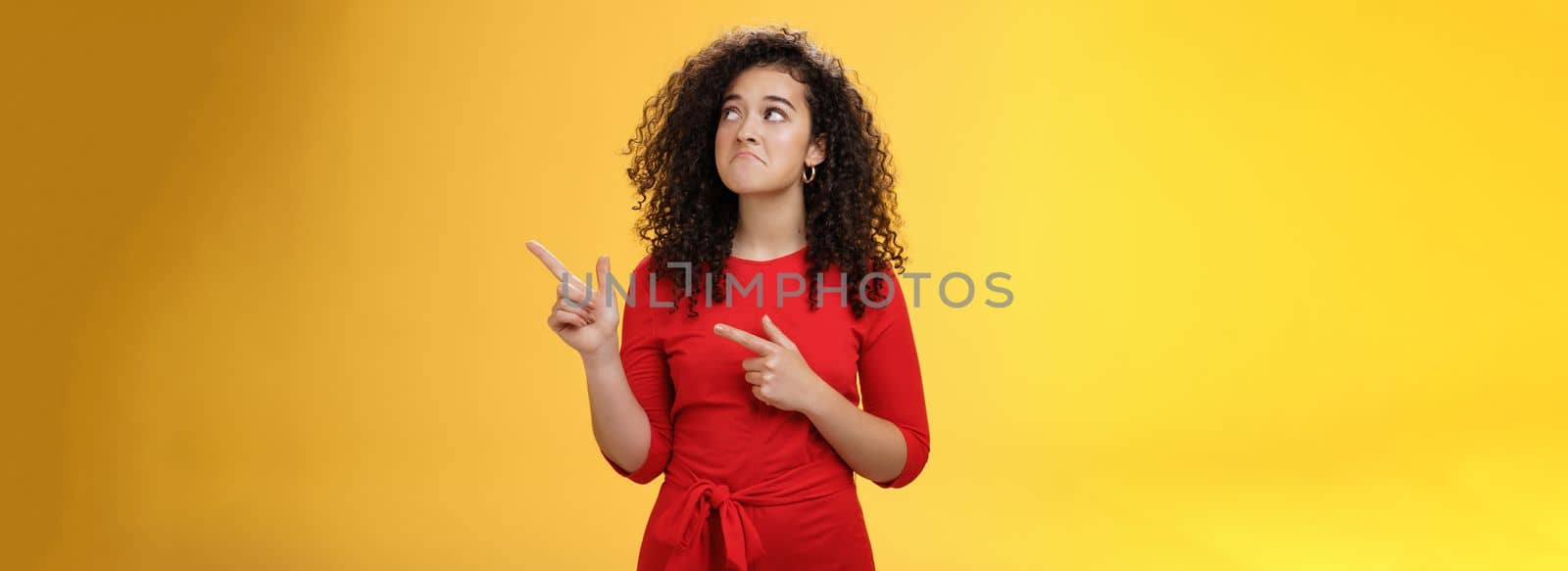 Lifestyle. Girl pointing and looking at upper right corner with regret and sorrow missing good chance or opportunity getting upset and sad pulling gloomy smile with lips pursed down standing over yellow wall.