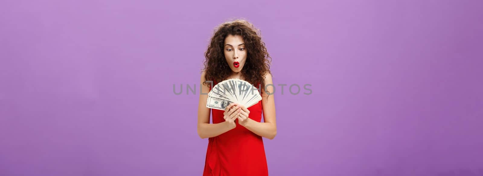 Portrait of surprised and amazed speechless woman folding lips making wow sound while holding and looking at lots of money being rich and successful receiving lots of cash over purple background. Finance concept