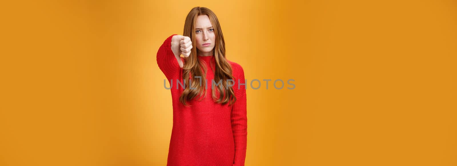 Disappointed upset and serious-looking attractive 20s redhead woman in red knitted dress frowning in dislike looking with judgement, showing thumb down in aversion and dissatisfaction over orange wall.