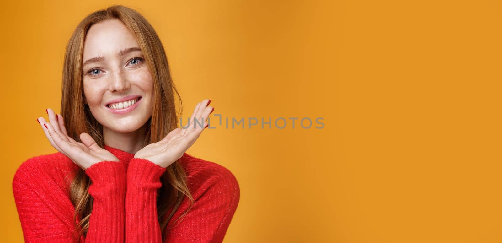 Close-up shot of sensual tender and gentle ginger girl. with romance and happy gaze holding palms near pure clean skin with freckles smiling joyfully tilting head looking friendly over orange wall.