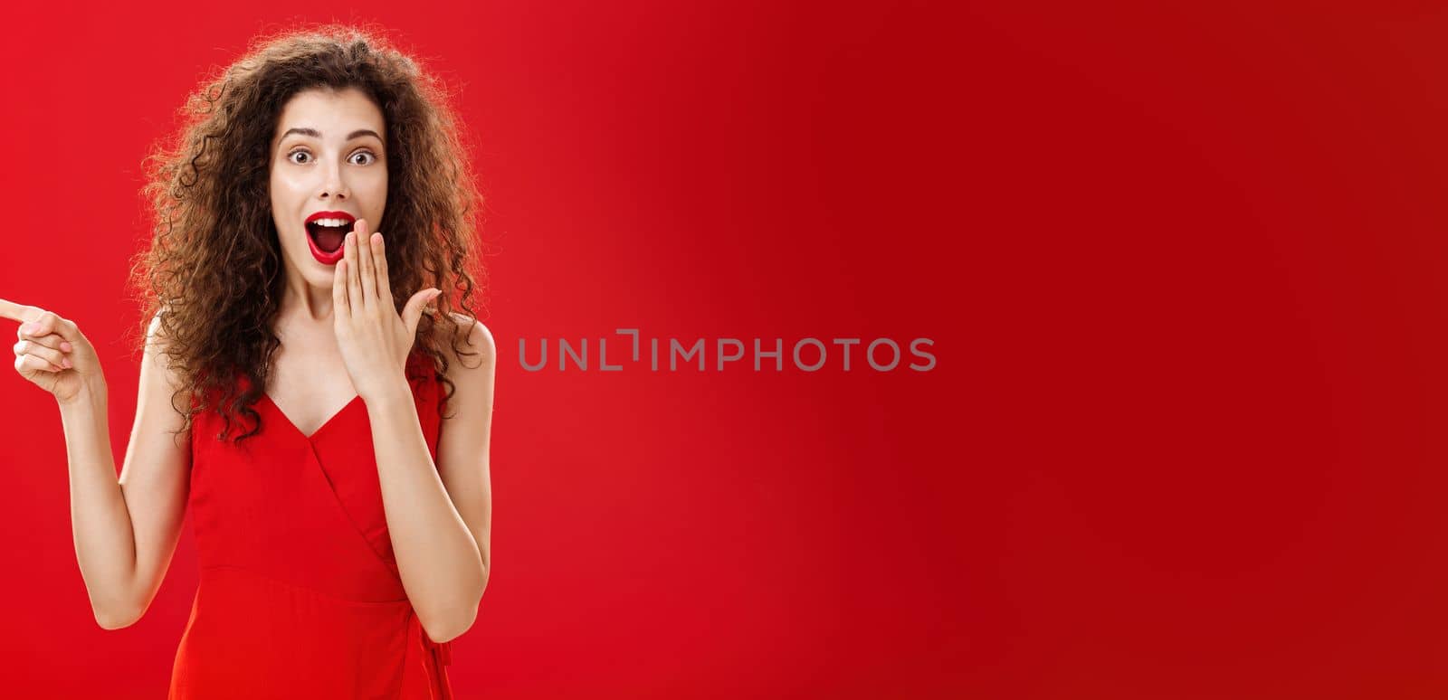 Happy surprised and delighted attractive curly-haired female in evening dress smiling amazed with opened mouth covering it with palm pointing left questioned and excited over red background. Emotions concept