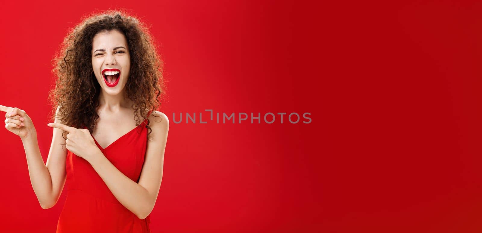Woman feeling awesome on great party of friend pointing at entrance. Stylish carefree curly-haired female in red evening dress having fun near pool winking smiling joyfully and pointing left.