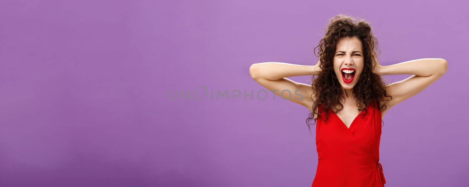 Ex-boyfriend spoilt woman formal event being outraged. and pissed yelling in fury holding hands on curly hair grimacing from anger standing over purple background in stylish evening red dress.