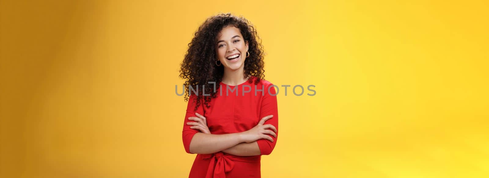 Self-assured happy enthusiastic curly-haired female reporter in cute red dress laughing carefree, having fun tilting head amused and holding hands crossed over body in confident pose over yellow wall.