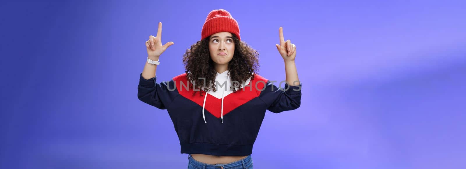 Girl feeling disappointed and gloomy as looking at bad weather making sad smile, pursing lips down raising hands staring up upset and unsatisfied standing displeased against blue background.