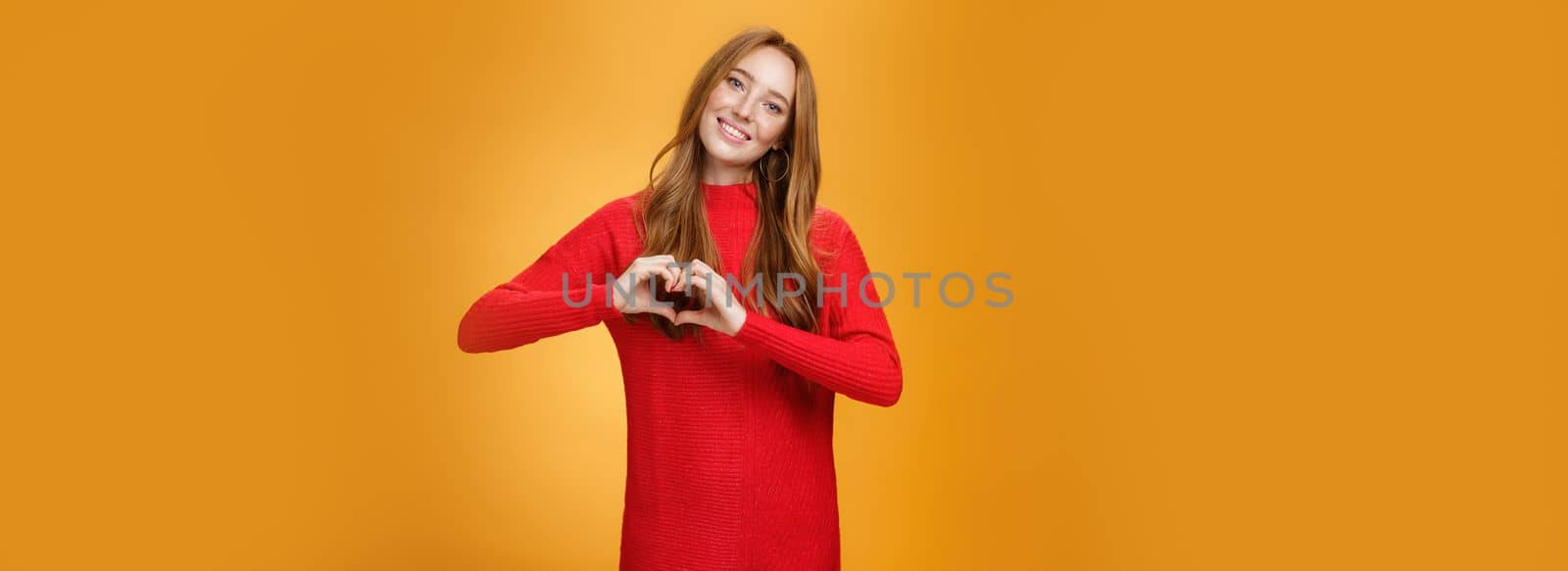 Attractive confident and cute ginger girl with freckles in red knitted warm dress showing love gesture tilting head and smiling broadly at camera liking and adoring new outfit over orange background by Benzoix