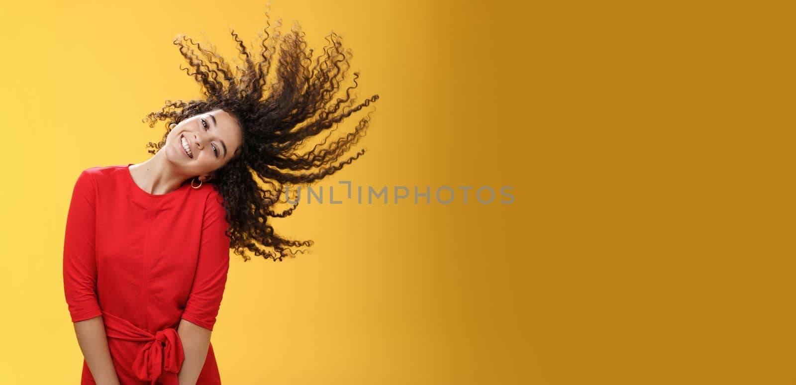Bright happy and carefree playful woman waving curly hair making wave and smiling broadly as standing joyful in red dress over yellow background in good mood for future adventures. Copy space