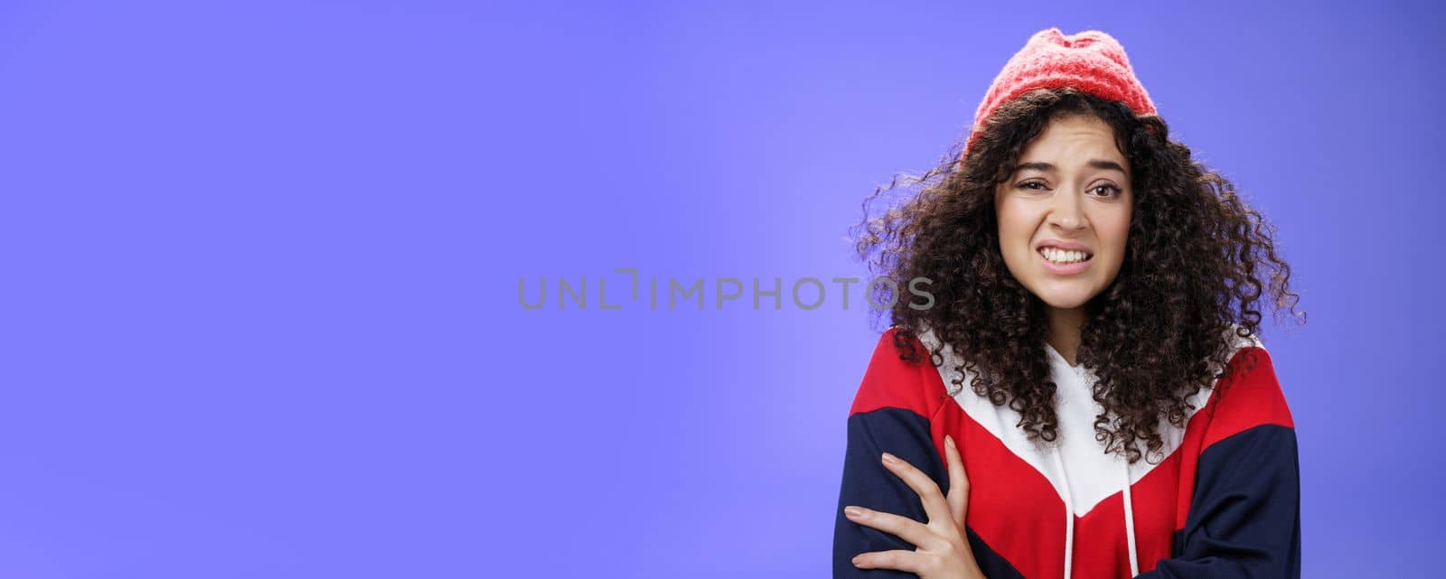 Woman with curly hair in winter beanie feeling uncomfortable and discomfort clenching teeth and frowning intense as hugging herself insecure and awkward, unwilling to say cruel rejection, feel awkward.