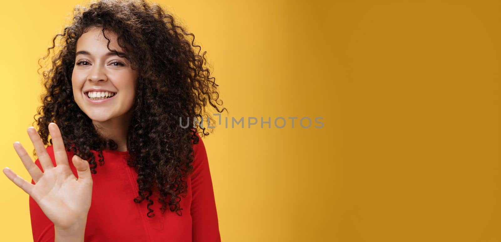 Lifestyle. Charming friendly and self-assured attractive curly woman waving cute with palm to say hi or hello smiling broadly greeting man trying flirt in party posing joyful over yellow background.