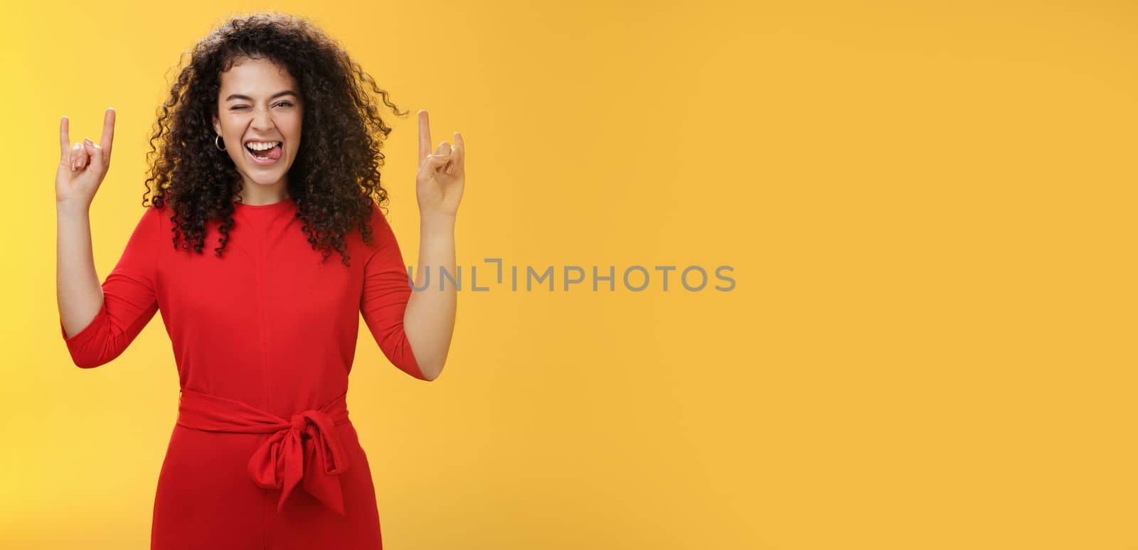 Cute excied and daring woman in 25s sticking out tongue as fooling around having fun on concert winking joyfully and showing rock-n-roll gesture feeling amused and alive over yellow background by Benzoix