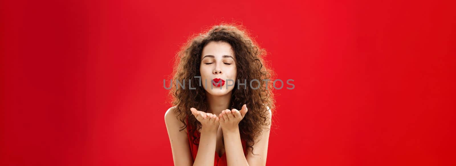 Tender and gentle stylish caucasian girl with curly hairstyle. and red lipstick bending towards camera with slight smile closed eyes and palms near folded lips blowing kiss at camera romantically.