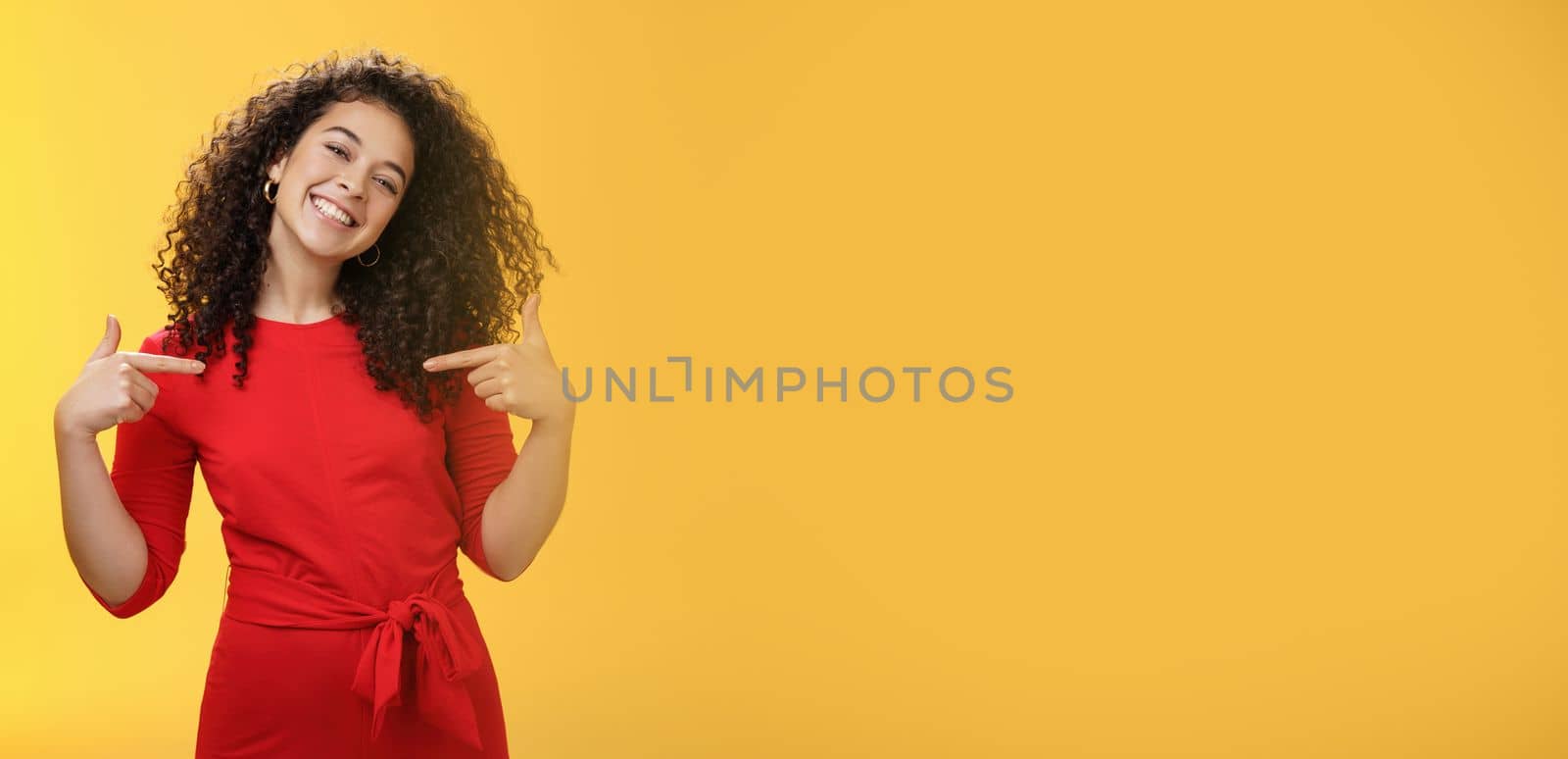 Lifestyle. Proud and satisfied ambitious successful female student in red dress standing pleased smiling and pointing at herself as if bragging about own achievements happily and glad over yellow wall.