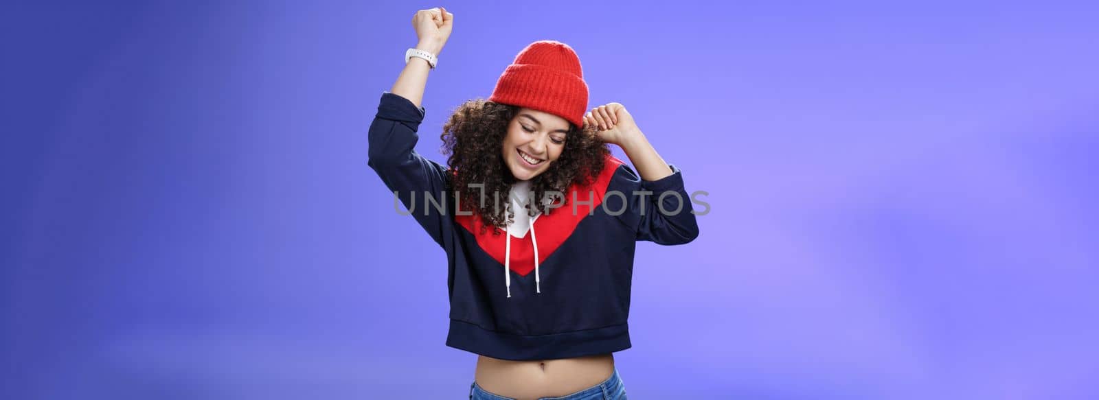 Girl celebrating favorite season in year. Portrait of carefree and joyful dancing woman with curly hair in cute red hat lifting hands in dance movements smiling enjoying music and holidays by Benzoix