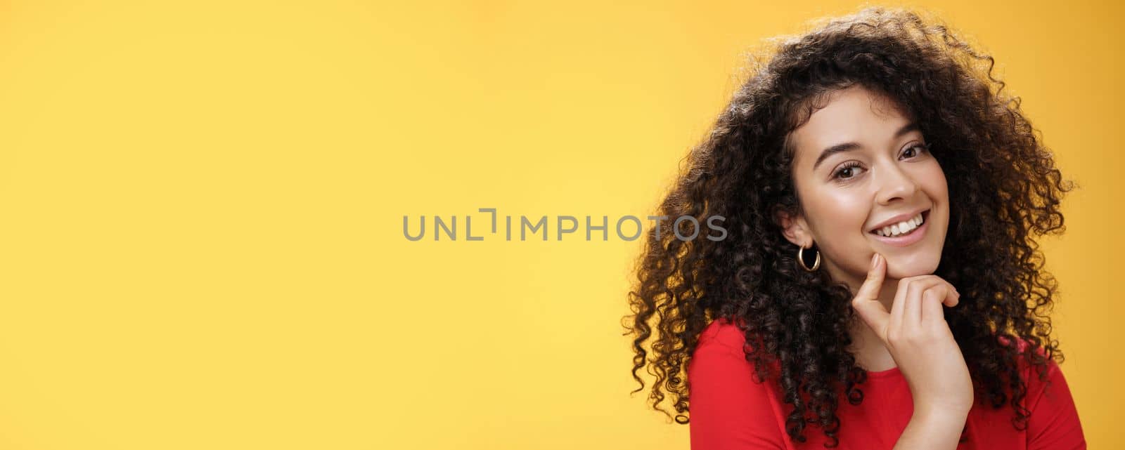 Headshot of cute silly and tender feminine romantic woman with curly hairstyle touching lip with index finger making eyes at camera and smiling as using seduction skills over yellow background by Benzoix